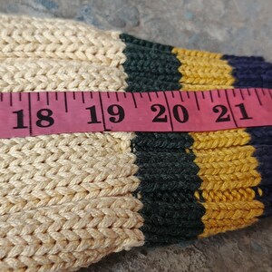 May include: Close-up of a cream-colored knitted sleeve with a pink measuring tape. The sleeve has green, yellow, and navy blue stripes at the cuff. The tape measures from 17 to 24 inches.