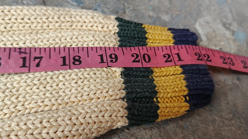 May include: Close-up of a cream-colored knitted sleeve with a pink measuring tape. The sleeve has green, yellow, and navy blue stripes at the cuff. The tape measures from 17 to 24 inches.