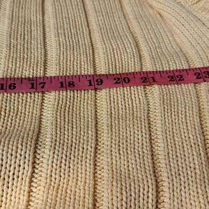 May include: Close-up of a light yellow knit sweater with a dark green accent. A pink measuring tape shows a length of approximately 13 to 26 inches. The sweater has a ribbed texture.