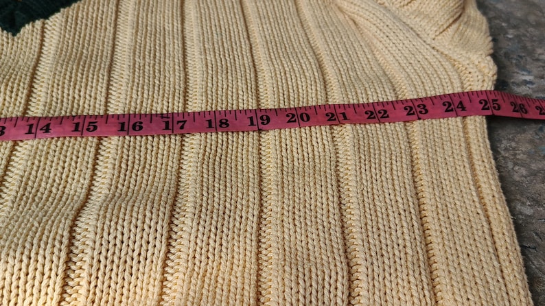 May include: Close-up of a light yellow knit sweater with a dark green accent. A pink measuring tape shows a length of approximately 13 to 26 inches. The sweater has a ribbed texture.