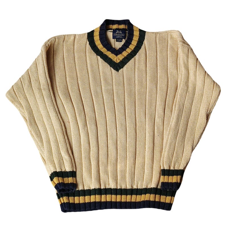 May include: Cream-colored ribbed knit sweater with a V-neck. The collar, cuffs, and hem feature navy blue, gold, and green stripes. The sweater has a relaxed fit and long sleeves.