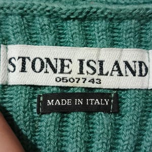 May include: Close-up of a teal knit fabric with a white label that reads "STONE ISLAND" and the number "0507743". Below the label is a black tag that says "MADE IN ITALY".