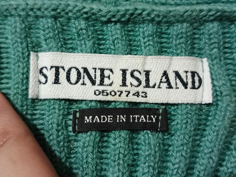 May include: Close-up of a teal knit fabric with a white label that reads "STONE ISLAND" and the number "0507743". Below the label is a black tag that says "MADE IN ITALY".
