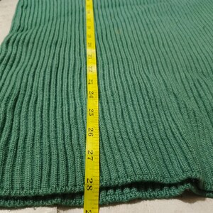May include: Close-up of a green knitted fabric with vertical ribbing. A yellow measuring tape is placed vertically along the fabric, showing a length of approximately 28 inches. The fabric appears to be a section of a garment.
