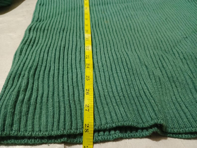 May include: Close-up of a green knitted fabric with vertical ribbing. A yellow measuring tape is placed vertically along the fabric, showing a length of approximately 28 inches. The fabric appears to be a section of a garment.