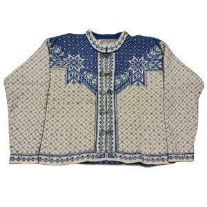 Op de afbeelding: Een crème, blauw en grijs patroon vest met een sneeuwvlok ontwerp. Het vest heeft een blauwe pas en een ruitpatroon op het lichaam. Het heeft een rij zilveren sluitingen aan de voorkant.