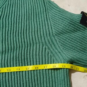 May include: Close-up of a teal green ribbed knit sweater. A yellow measuring tape shows a length of approximately 11 to 23 inches. A black patch with a gold emblem is on the upper sleeve.