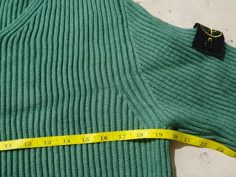May include: Close-up of a teal green ribbed knit sweater. A yellow measuring tape shows a length of approximately 11 to 23 inches. A black patch with a gold emblem is on the upper sleeve.