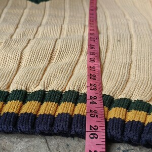 May include: Cream-colored knit sweater with ribbed design and a V-neck. The sweater has a striped pattern in navy blue, gold, and green at the bottom. A pink measuring tape shows a length of approximately 26 inches.