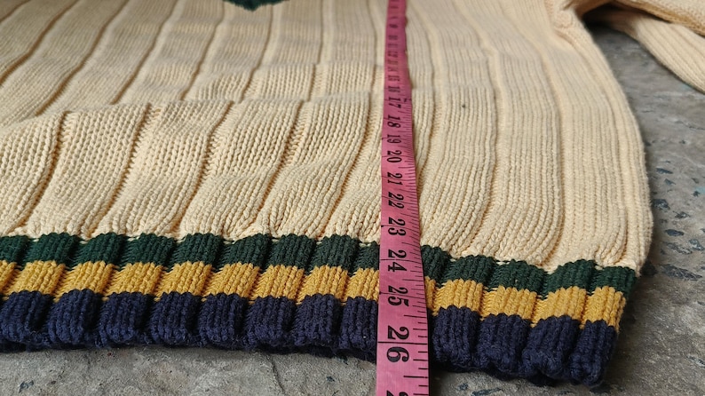 May include: Cream-colored knit sweater with ribbed design and a V-neck. The sweater has a striped pattern in navy blue, gold, and green at the bottom. A pink measuring tape shows a length of approximately 26 inches.