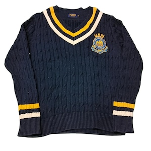 May include: Navy blue cable knit sweater with a V-neck collar. The collar and cuffs feature yellow and white stripes. A crest with a crown and eagle is on the chest. The brand name "PLORY" is visible on the collar.