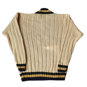 May include: Cream-colored knit sweater with vertical ribbing. The collar and cuffs feature navy blue, green, and gold stripes. The sweater has a relaxed fit and a classic, vintage style.