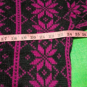 May include: Close-up of a black and fuchsia sweater with a snowflake pattern. A pink measuring tape is laid across the sweater, showing measurements from 16 to 28 inches. The sweater has a knitted texture.