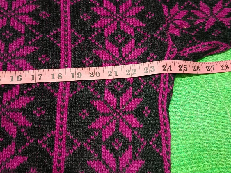 May include: Close-up of a black and fuchsia sweater with a snowflake pattern. A pink measuring tape is laid across the sweater, showing measurements from 16 to 28 inches. The sweater has a knitted texture.