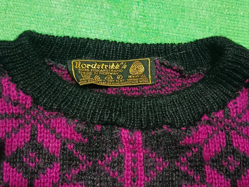 May include: Close-up of a vintage Nordstrikk sweater. The sweater features a black crew neck and a pattern of magenta and black geometric shapes. A tag with the brand name and origin is visible.