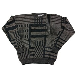 May include: A gray and black patterned sweater with a crew neck and long sleeves. The sweater features a patchwork design with various geometric shapes and textures. The cuffs and hem are ribbed. The sweater is made of a soft, textured material.
