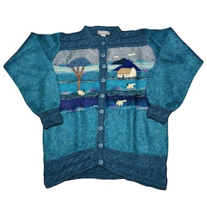 Könnte beinhalten: Türkisblaue Strickjacke mit Landschaftsdesign. Der Cardigan mit Knöpfen zeigt eine Szene mit Baum, Haus und Schafen. Der Pullover hat lange Ärmel und ein Zopfmuster am unteren Saum.