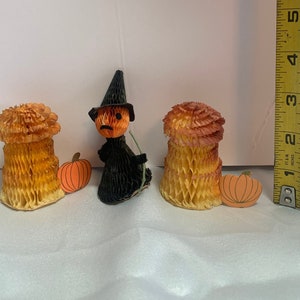 May include: Three Halloween decorations made of paper. A witch with a black hat and orange face holds a broom. Two orange pumpkins with black stems are next to the witch. Two orange and yellow honeycomb decorations are on either side of the witch.