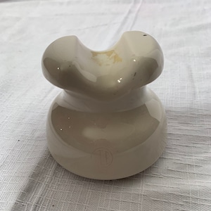May include: A white ceramic insulator with a rounded top and a flat base. The insulator has a small hole in the top and a circular mark on the base.