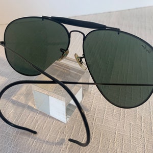 May include: Black aviator sunglasses with green lenses and a black metal frame. The sunglasses are on a clear display stand. The sunglasses have a Ray-Ban logo on the right lens.