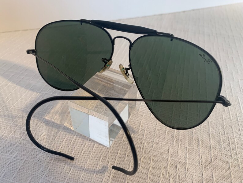 May include: Black aviator sunglasses with green lenses and a black metal frame. The sunglasses are on a clear display stand. The sunglasses have a Ray-Ban logo on the right lens.