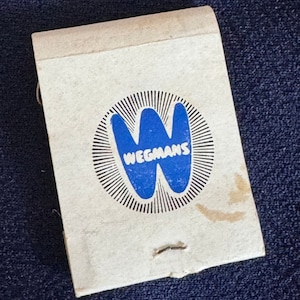 May include: A white matchbook with a blue and black logo. The logo is a stylized "W" with the word "Wegmans" underneath.