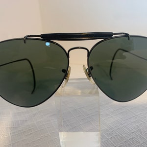 May include: Black Ray-Ban aviator sunglasses with green lenses. The sunglasses are on a white surface.