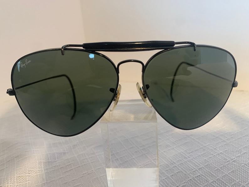 May include: Black Ray-Ban aviator sunglasses with green lenses. The sunglasses are on a white surface.