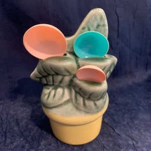 May include: A ceramic planter shaped like a plant with three leaves. The planter has three colorful measuring spoons in the leaves. The planter is yellow and the leaves are green.
