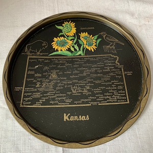 May include: A black metal tray with a gold rim features a map of Kansas with the names of cities and towns. The tray also has a sunflower design and the words "Sunflower" and "Kansas".
