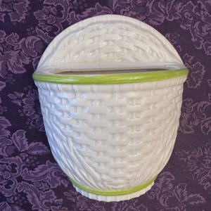 May include: White ceramic wall basket with a woven texture. The basket has a curved top and a green trim around the opening and base. The basket is designed to be hung on a wall and is suitable for displaying plants or other decorative items.