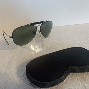 May include: Black Ray-Ban aviator sunglasses with green lenses and a metal frame. The sunglasses are on a clear display stand and come with a black carrying case.