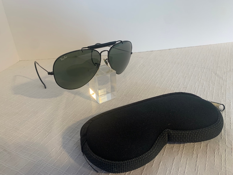 May include: Black Ray-Ban aviator sunglasses with green lenses and a metal frame. The sunglasses are on a clear display stand and come with a black carrying case.