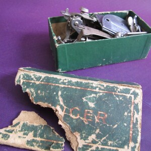 May include: A green cardboard box with a torn lid, revealing a collection of vintage sewing machine attachments. The box is labeled "CER" in gold lettering.