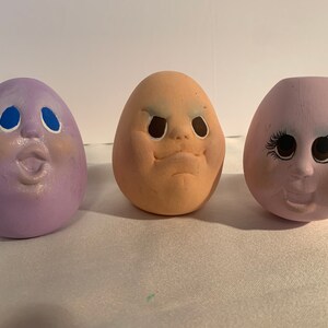 May include: Three ceramic egg-shaped ornaments with painted faces. The first ornament is purple with blue eyes and a surprised expression. The second ornament is peach with brown eyes and a frowning expression. The third ornament is purple with brown eyes and a smiling expression.