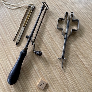 May include: Three antique kitchen tools are displayed on a wooden surface. The tools include a brass and steel needle, a black-handled peeler with a crank, and a wire whisk. A Scrabble tile with the letter "F" and the number "4" is also present.