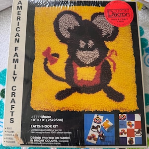 May include: An American Family Crafts latch hook kit #111-Mouse, measuring 10 inches by 10 inches. The packaging features a yellow background with a black mouse graphic, a red apron, and a red bow. Includes polyester or acrylic yarn and cotton canvas.