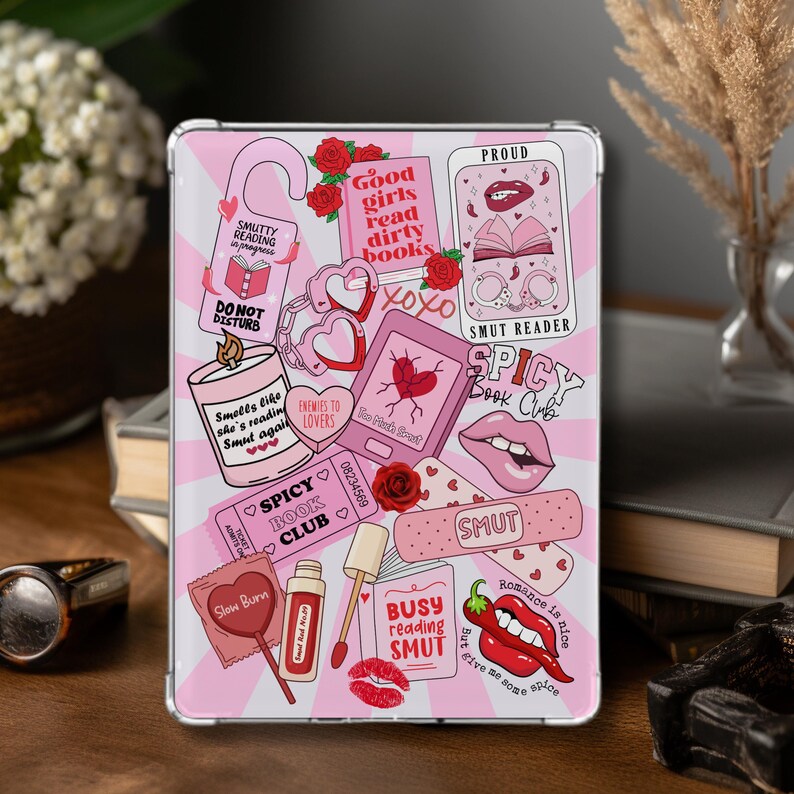 以下が含まれることがあります： ピンクと白の模様のタブレットケース。バラ、ハート、唇、そして「Smut Reader」や「Spicy Book Club」などのテキストを含む様々なステッカーとイラストが描かれています。