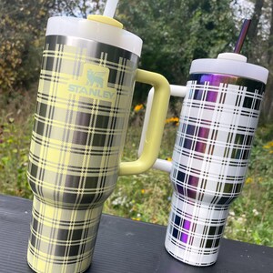 Könnte beinhalten: Zwei Stanley-Becher aus Edelstahl mit Karomustern. Einer hat ein gelb-schwarzes Karomuster mit gelbem Griff und dem Stanley-Logo. Der andere hat ein weiß-schwarzes Karomuster mit einem violett-blauen, schillernden Finish.