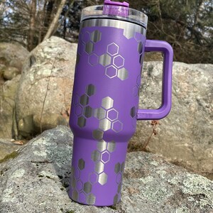 May include: A purple stainless steel travel mug with a honeycomb pattern and a lid. The mug has a handle and is sitting on a rock.