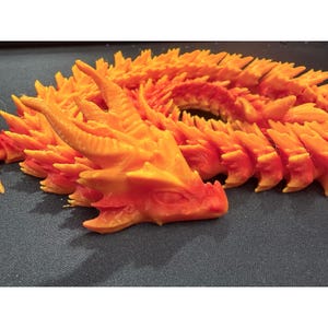 May include: A 3D printed articulated dragon figure featuring a vibrant gradient of orange and red colors. The creature has a detailed head with prominent horns and a long, segmented body with sharp, spiky scales that curve across the frame.