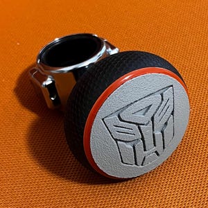 May include: A black and gray car gear shift knob with an orange trim and a gray Transformers logo.