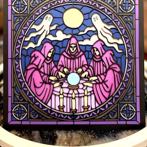 May include: A layered, square art piece depicting a stained-glass window scene. The design features figures in pink robes around a table with a crystal ball, candles, and ghosts. The background is blue with a yellow moon.