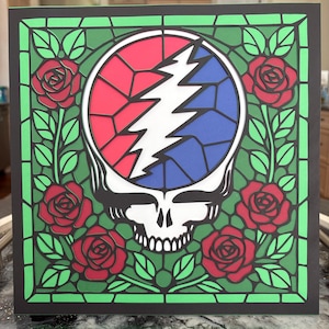 Może przedstawiać: Wielowarstwowe dzieło sztuki papierowej przedstawiające czaszkę Steal Your Face z czerwonymi, niebieskimi i białymi akcentami. Projekt otoczony jest czerwonymi różami i zielonymi liśćmi na czarno-zielonym tle.