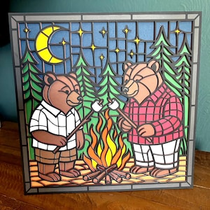 May include: A layered wood art piece depicting two bears roasting marshmallows over a campfire. The bears are in a forest setting with a night sky, moon, and stars. One bear wears a white plaid shirt, the other a red plaid shirt. The artwork has a stained glass effect.