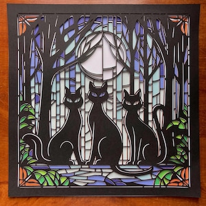 May include: A black paper cut art piece featuring three black cats sitting in front of a stained-glass window with a full moon. The background includes trees and colorful accents of blue, green, orange, and white.
