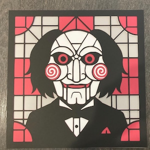 May include: Square print featuring a stylized illustration of the Jigsaw puppet from the Saw horror film series. The puppet's face is white with red eyes and spiral cheeks, set against a red and white stained-glass-style background.