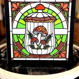 May include: A square, black-framed art print featuring a stained-glass style design. The artwork showcases a cluster of mushrooms within a cage-like structure, surrounded by floral and geometric elements in shades of orange, green, brown, and white.
