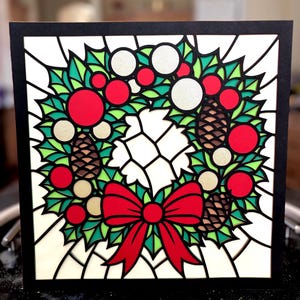 May include: A Christmas card featuring a stained-glass style wreath design. The wreath includes red berries, green leaves, pinecones, and a red bow. The card has a black border and a white background.