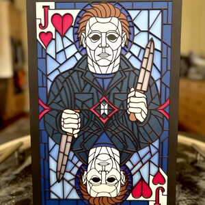 May include: A layered paper art piece depicting a figure in a stained-glass style, holding a knife. The design features a blue and white background, with the figure's face and hands in white. The card is framed in black, with the Jack of Hearts symbols.